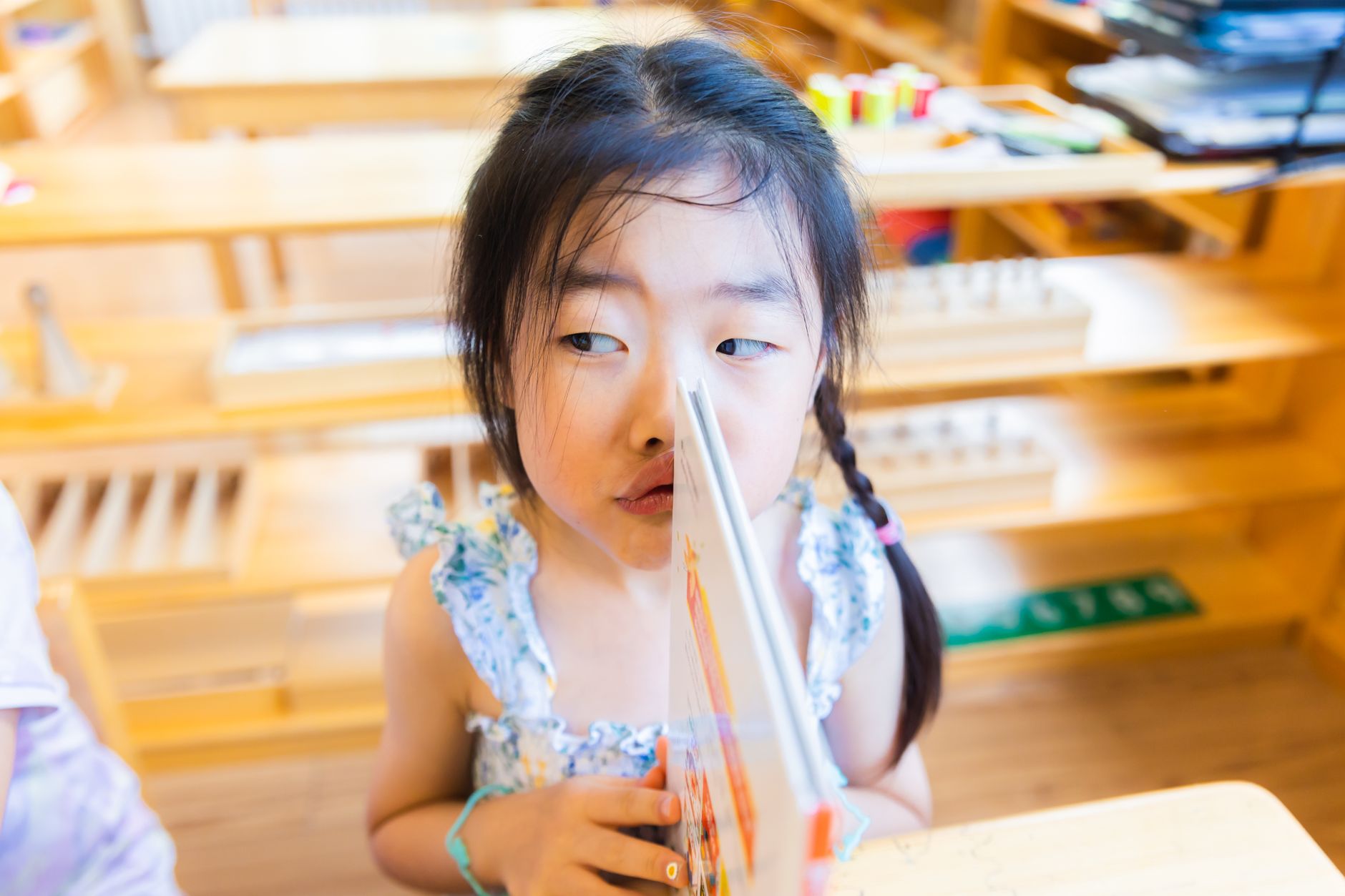 Cute girl playfully holding a book in a colorful elementary classroom.